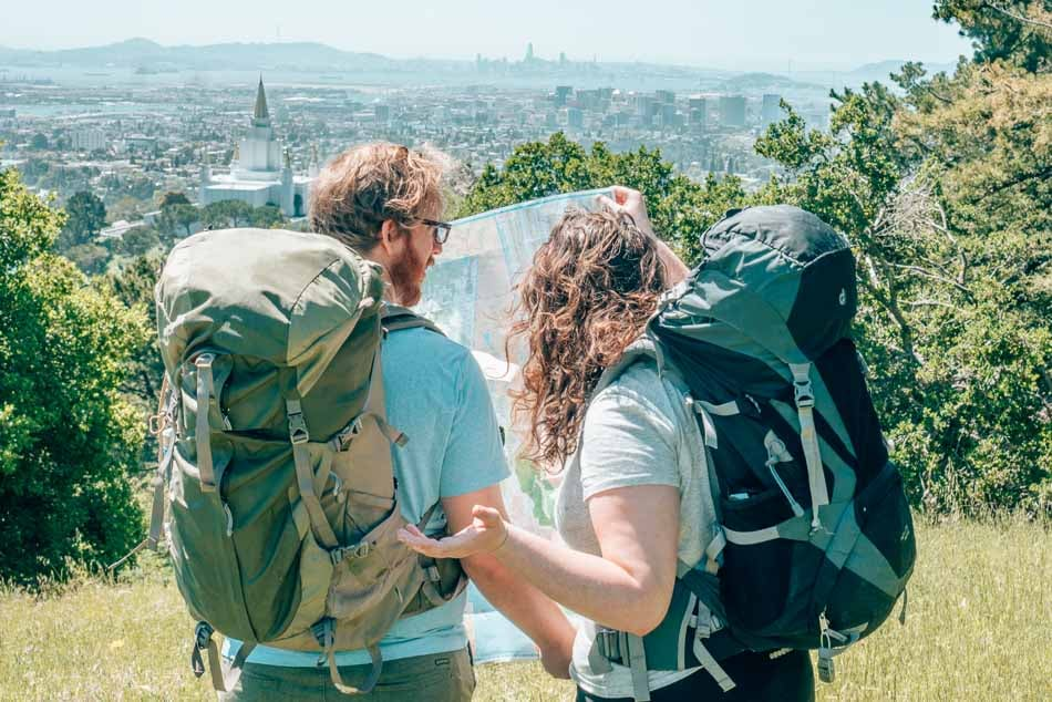 Young backpackers with large hiking backpacks consulting a map while overlooking a city, symbolizing budget travel and adventure