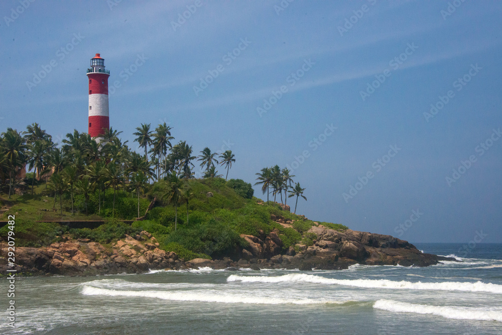The red and white Kovalam Beach lighthouse stands amidst palm trees, overlooking the Arabian Sea waves in Kerala